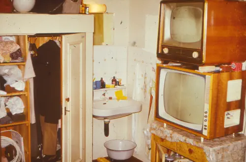 Kleiner geöffneter Kleiderschrank, daneben in der Ecke ein Waschbecken,
anscließend rechts davon zwei übereinander gestellte alte Röhrenfernseher auf
einer kleinen Kommode